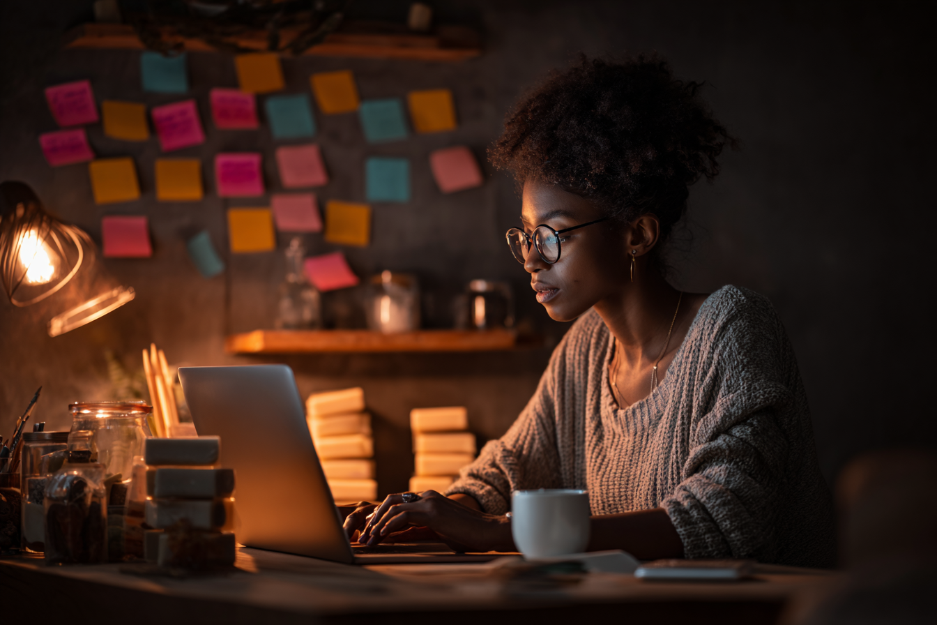 A young Nigerian woman working on her laptop in a cozy workspace, surrounded by packaged handmade products.